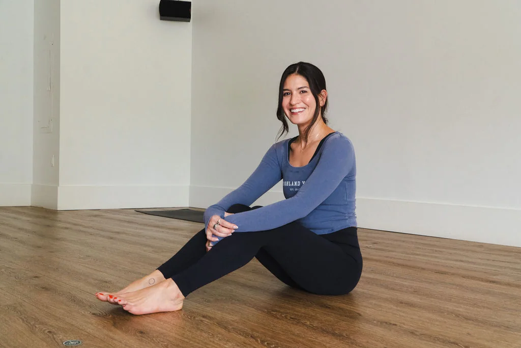 A person smiling while seated on the studio floor in a blue Highland Yoga long-sleeve top